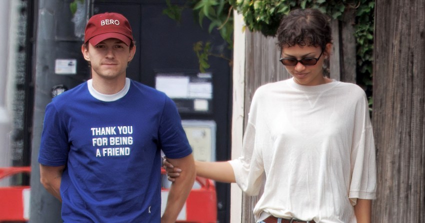 Man in blue 'Thank You For Being A Friend' T-shirt and red cap walks a French bulldog alongside a woman in a white tee and jeans on a London sidewalk.