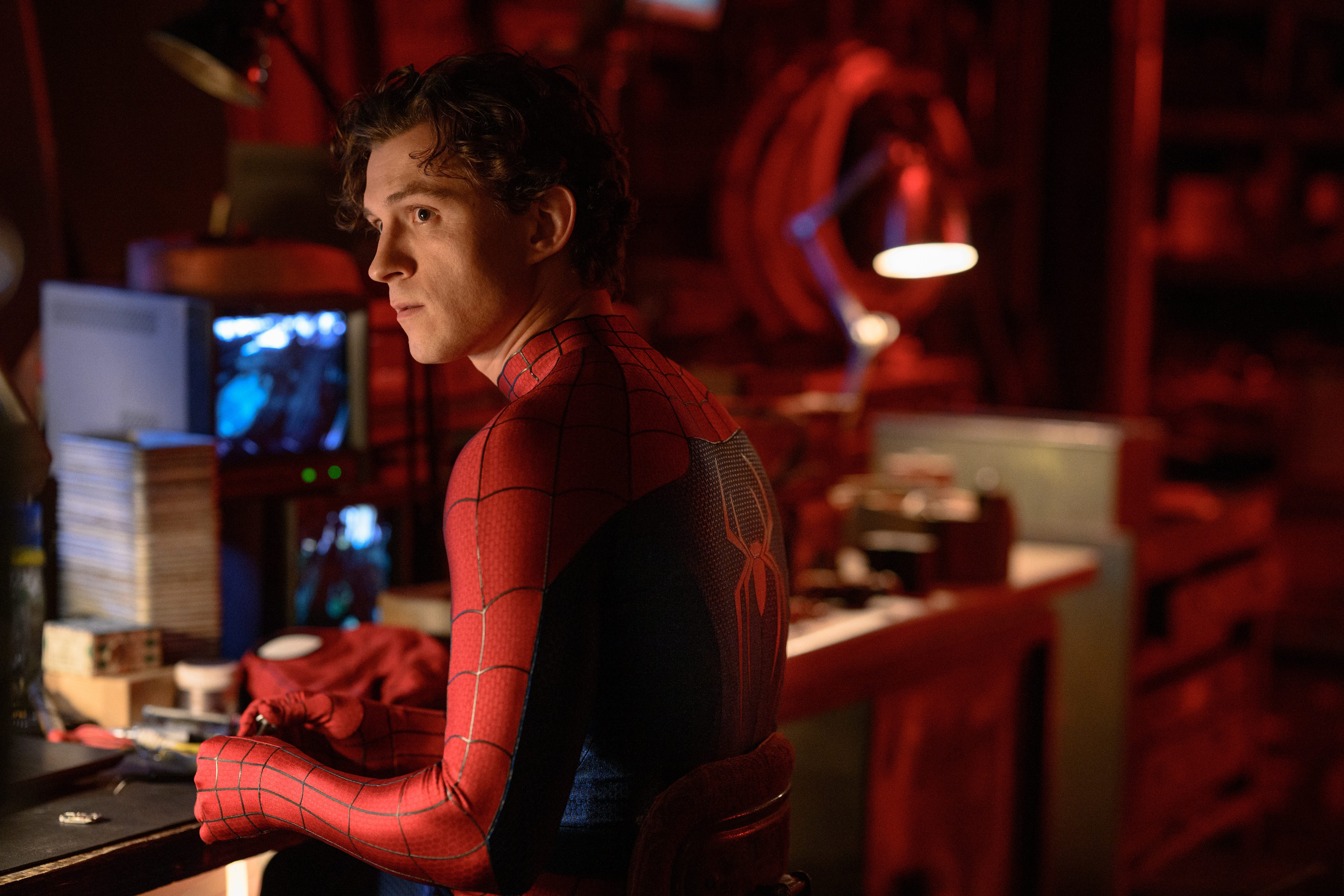 A young man in a red and black Spider-Man suit leans against a workbench in a red-lit workshop, looking to the side.