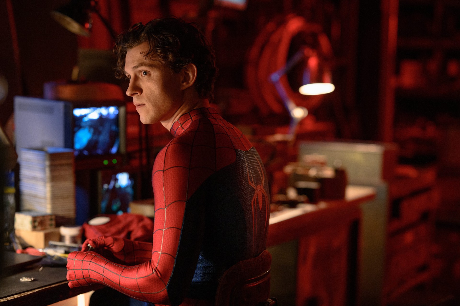 A young man in a red and black Spider-Man suit leans against a workbench in a red-lit workshop, looking to the side.