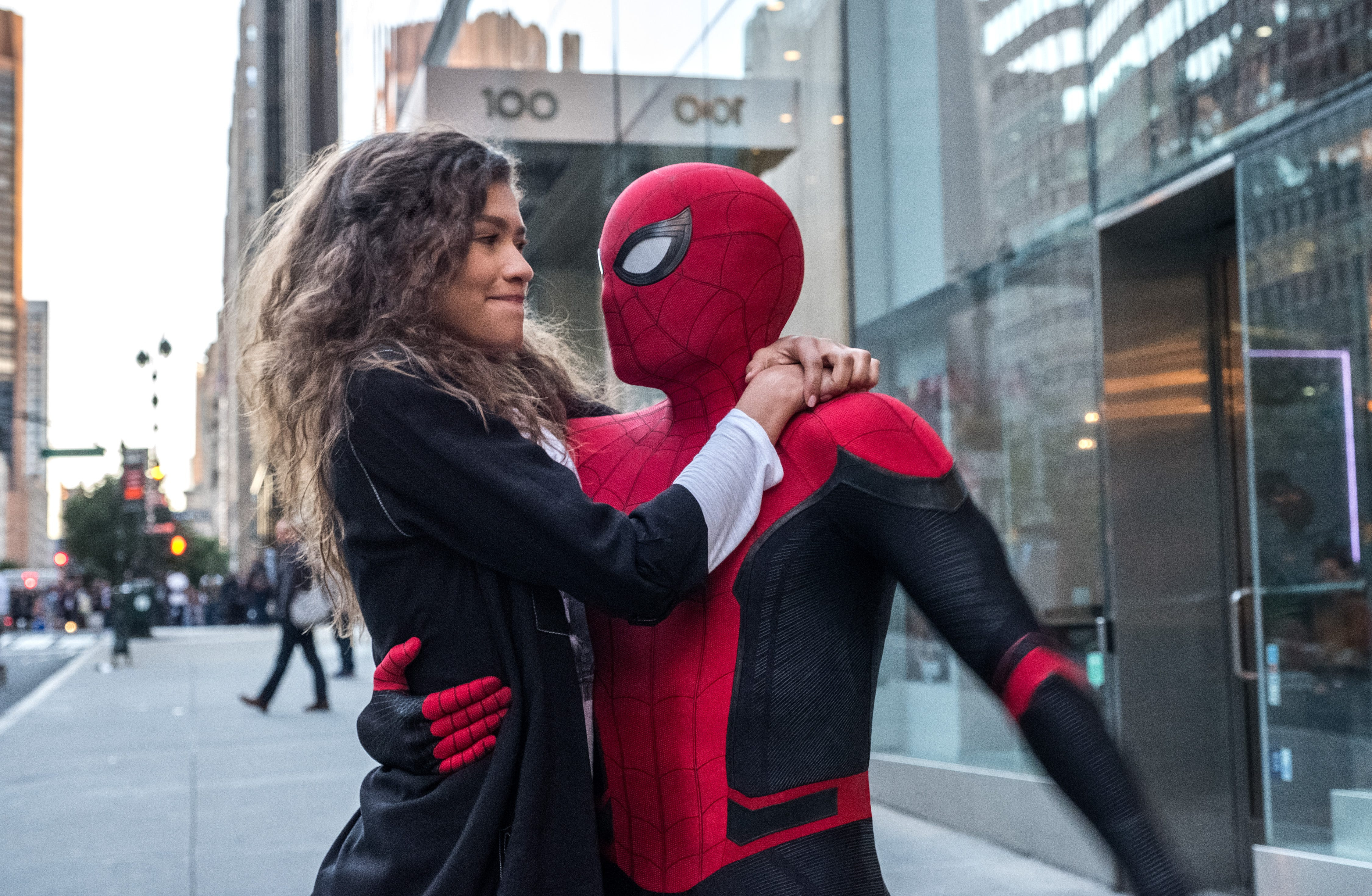 A young woman in a black jacket embraces a person in a red and black Spider-Man suit on a city sidewalk.