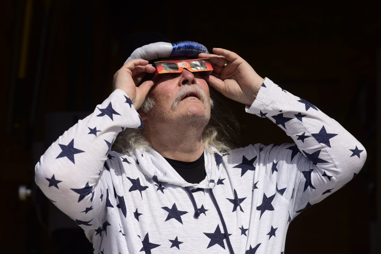 Man in a white star-print hoodie holding red-framed solar eclipse glasses to his eyes and looking up at the sky.