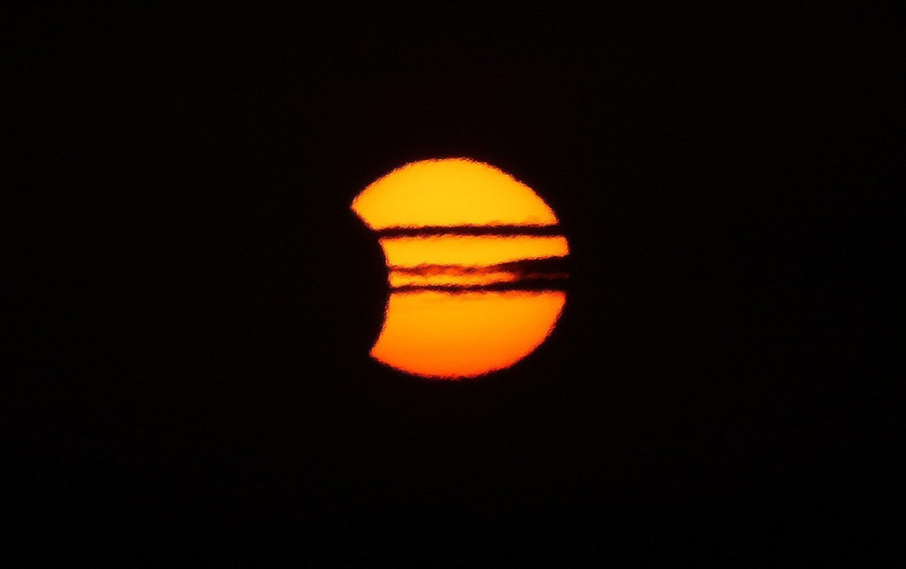 Close-up telescopic view of a partially eclipsed orange sun with two dark cloud bands crossing its face against a black background.