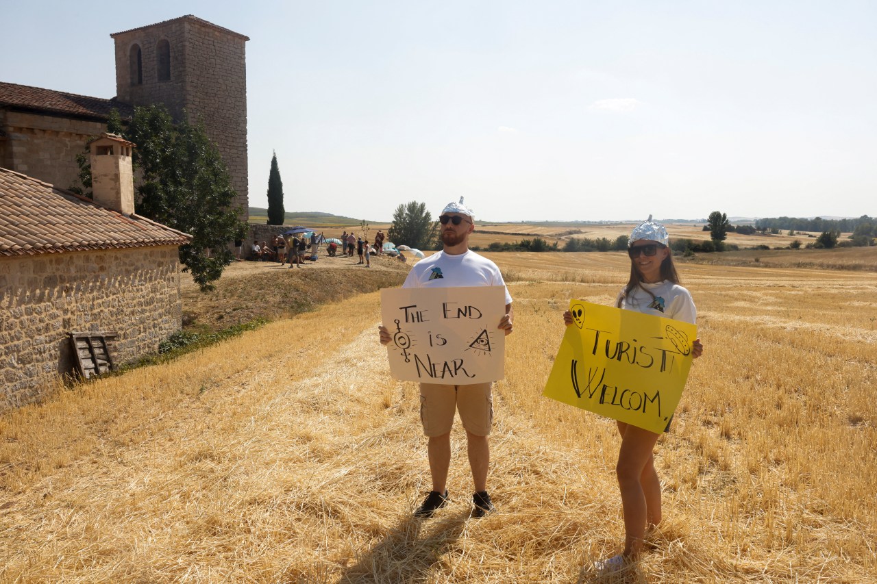 A man and woman wearing tinfoil hats stand in a dry wheat field near a stone church, holding signs reading 'The End Is Near' and 'Turisti Welcom' ahead of the solar eclipse in Villalibado, Spain.