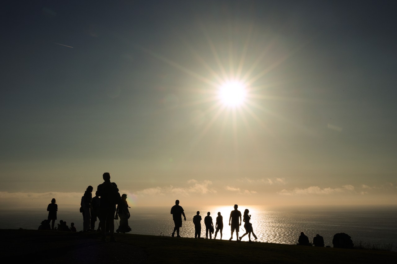 Silhouettes of a crowd of people standing on a hilltop overlooking the ocean, with a bright starburst sun low in the sky above them.