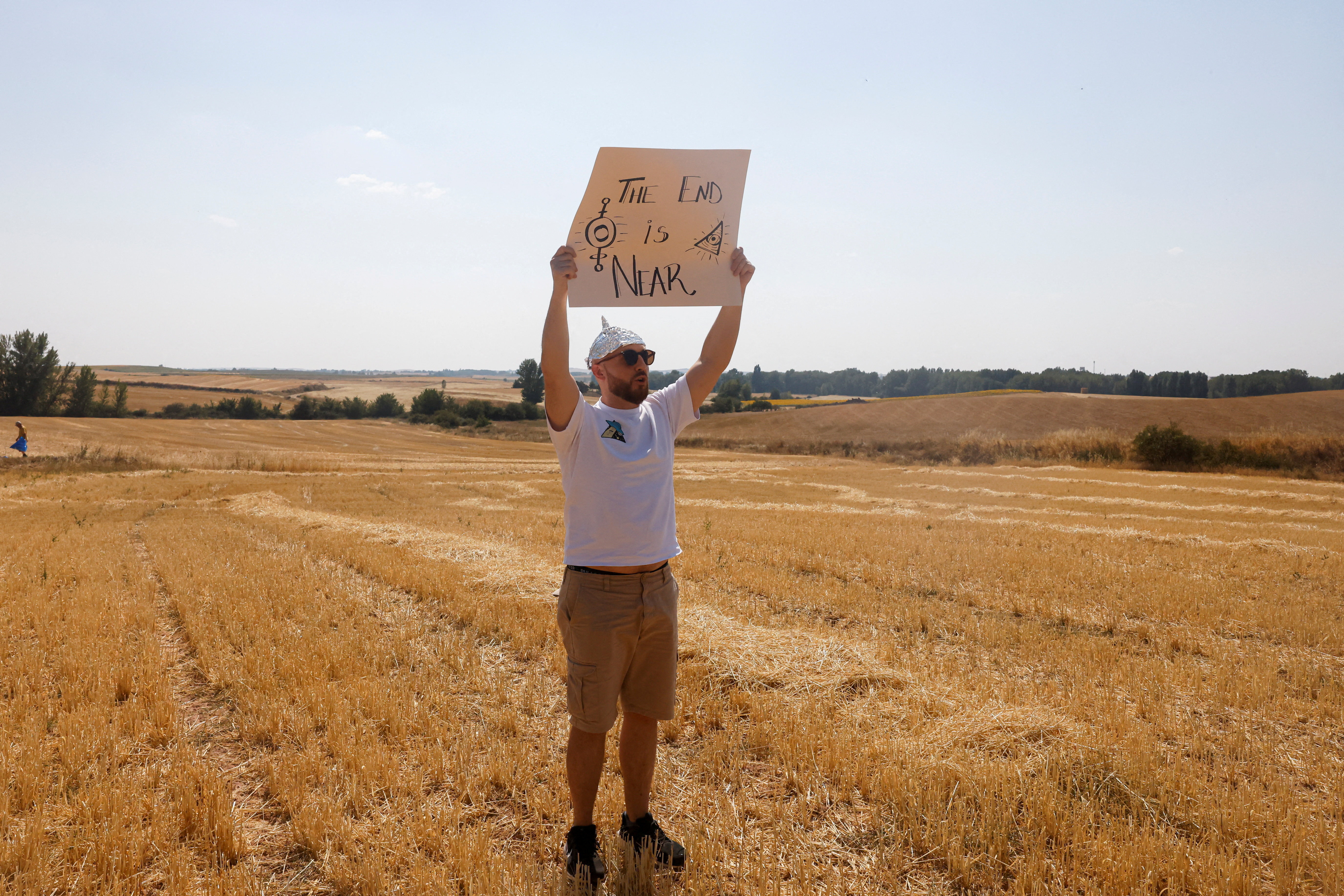 Man in a tinfoil hat and sunglasses holds a cardboard sign reading 'The End is Near' above his head while standing in a dry, golden field.