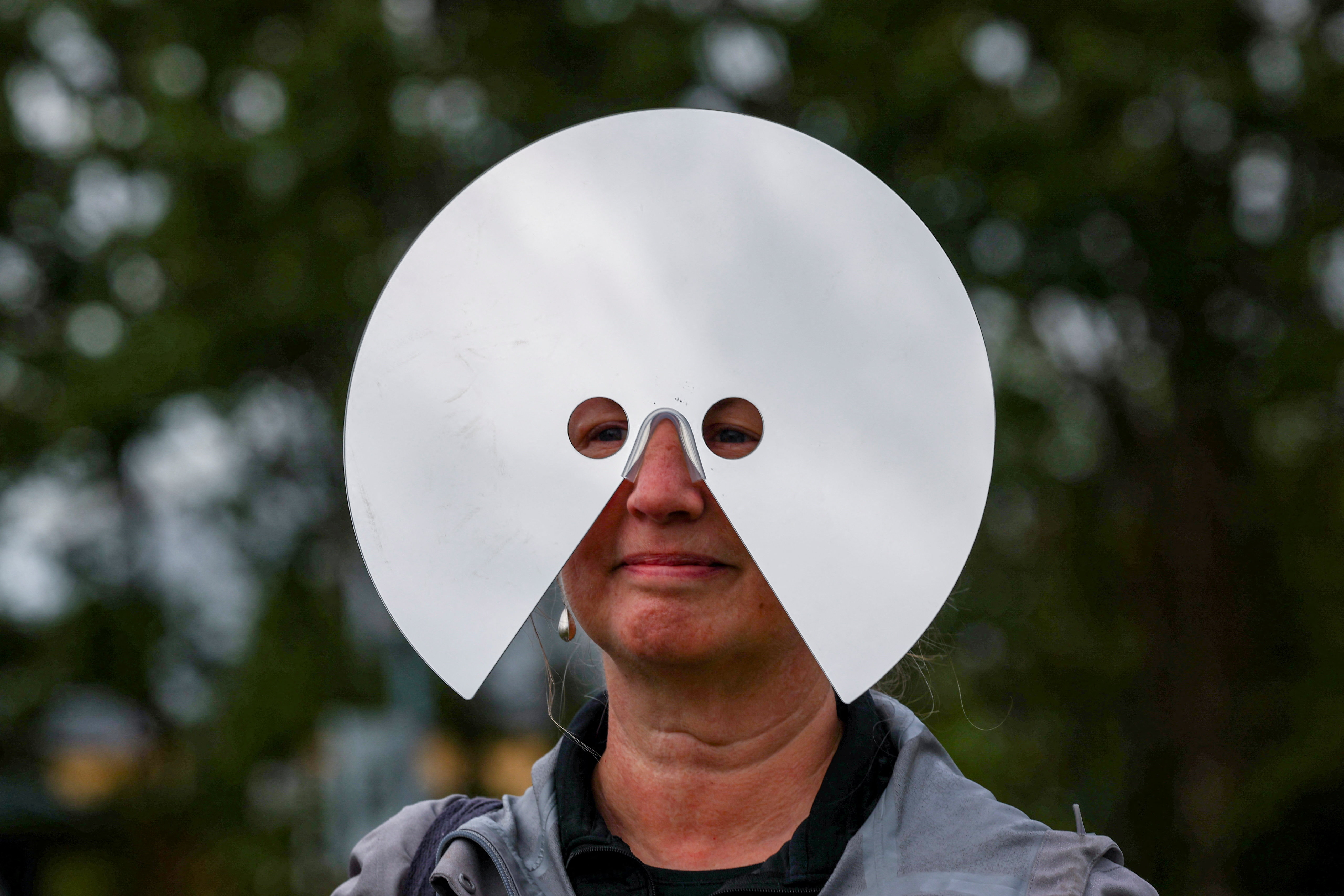 A woman wearing a large round silver face mask with two small eye holes looks toward the sky during a solar eclipse in Reykjavik, Iceland.