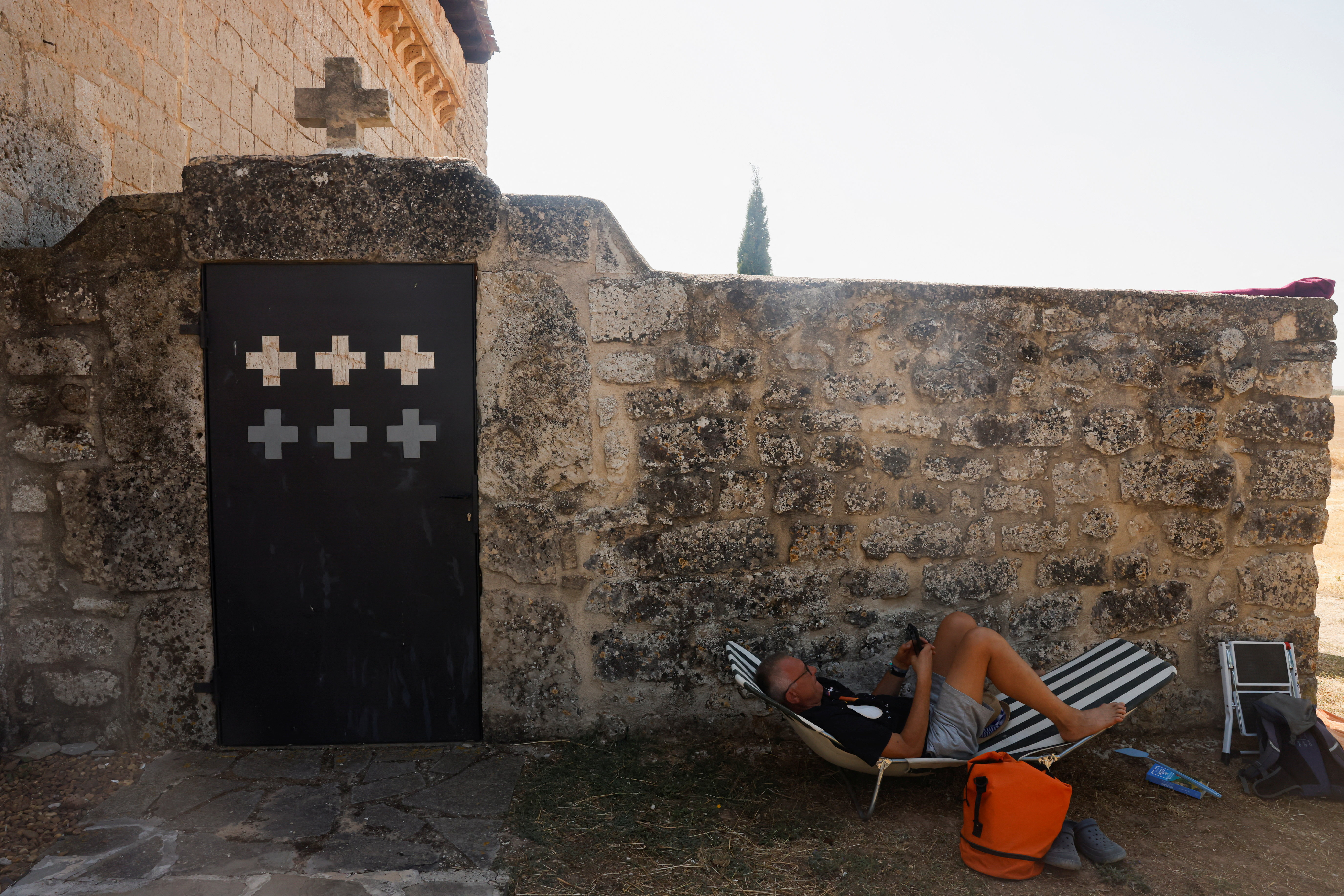 Man resting in a black-and-white striped folding chair leaning against a stone wall with a black door decorated with white crosses, bags at his feet.