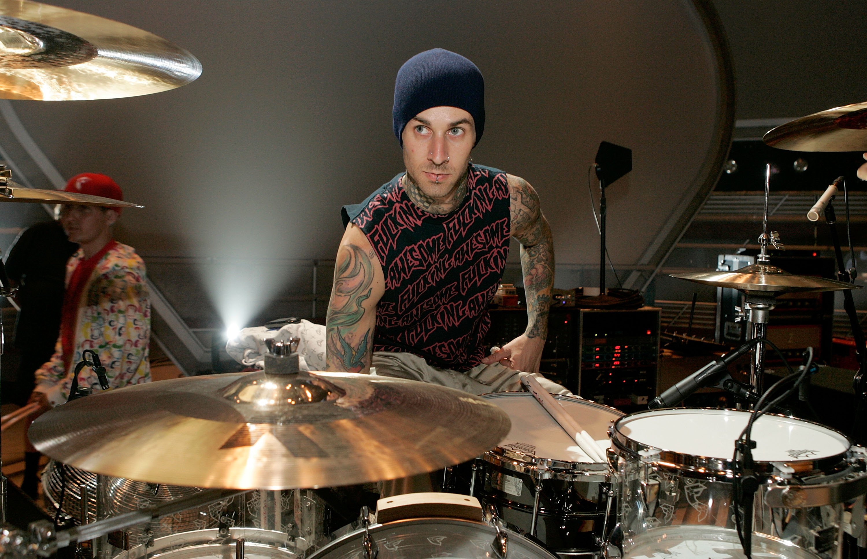 Tattooed man in a blue beanie and sleeveless shirt leaning over a drum kit surrounded by cymbals in a large arena rehearsal setting.