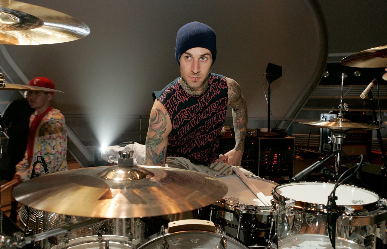 Tattooed man in a blue beanie and sleeveless shirt leaning over a drum kit surrounded by cymbals in a large arena rehearsal setting.