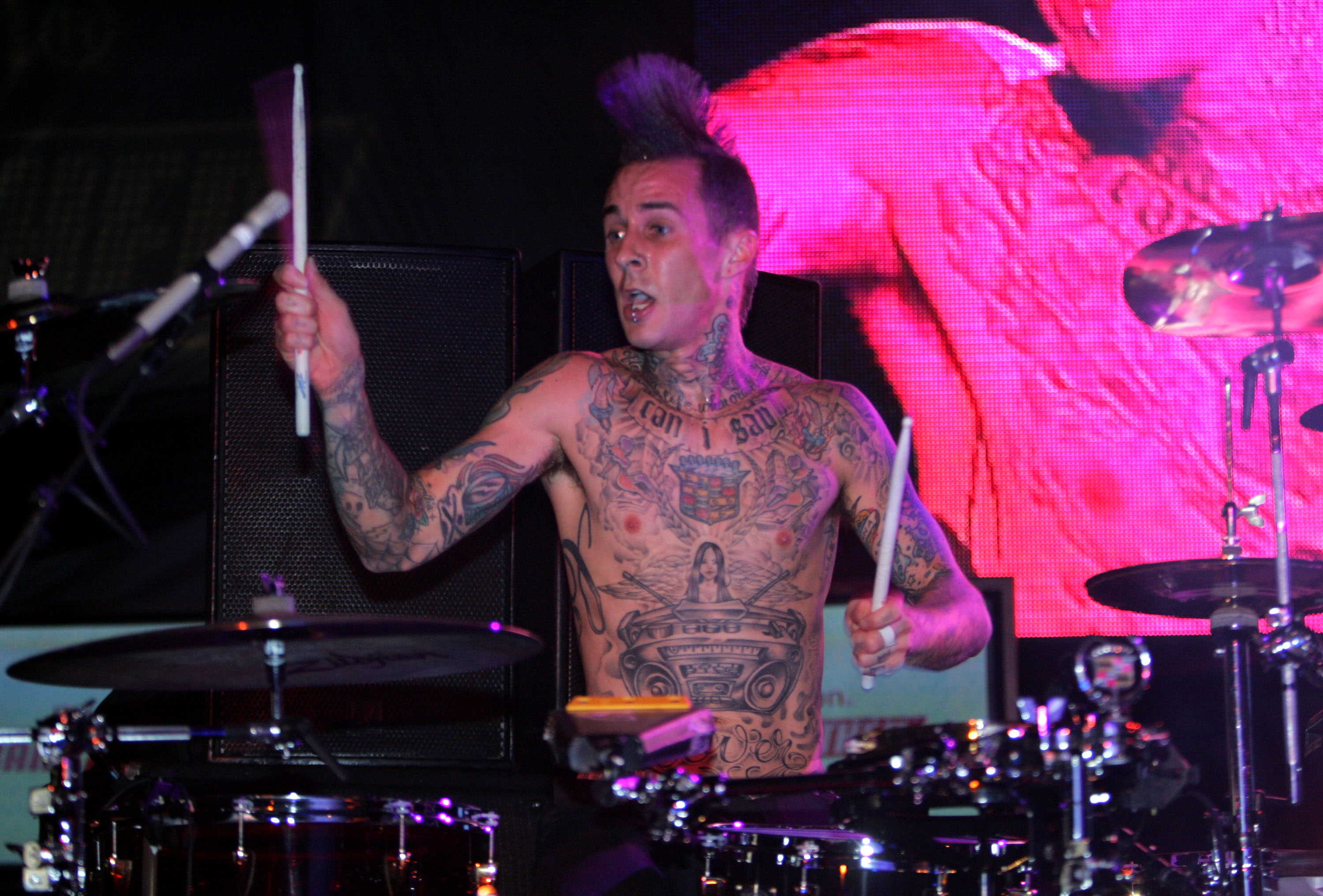 Shirtless tattooed drummer with a mohawk playing a drum kit on stage under pink and purple lights, drumsticks raised in both hands.