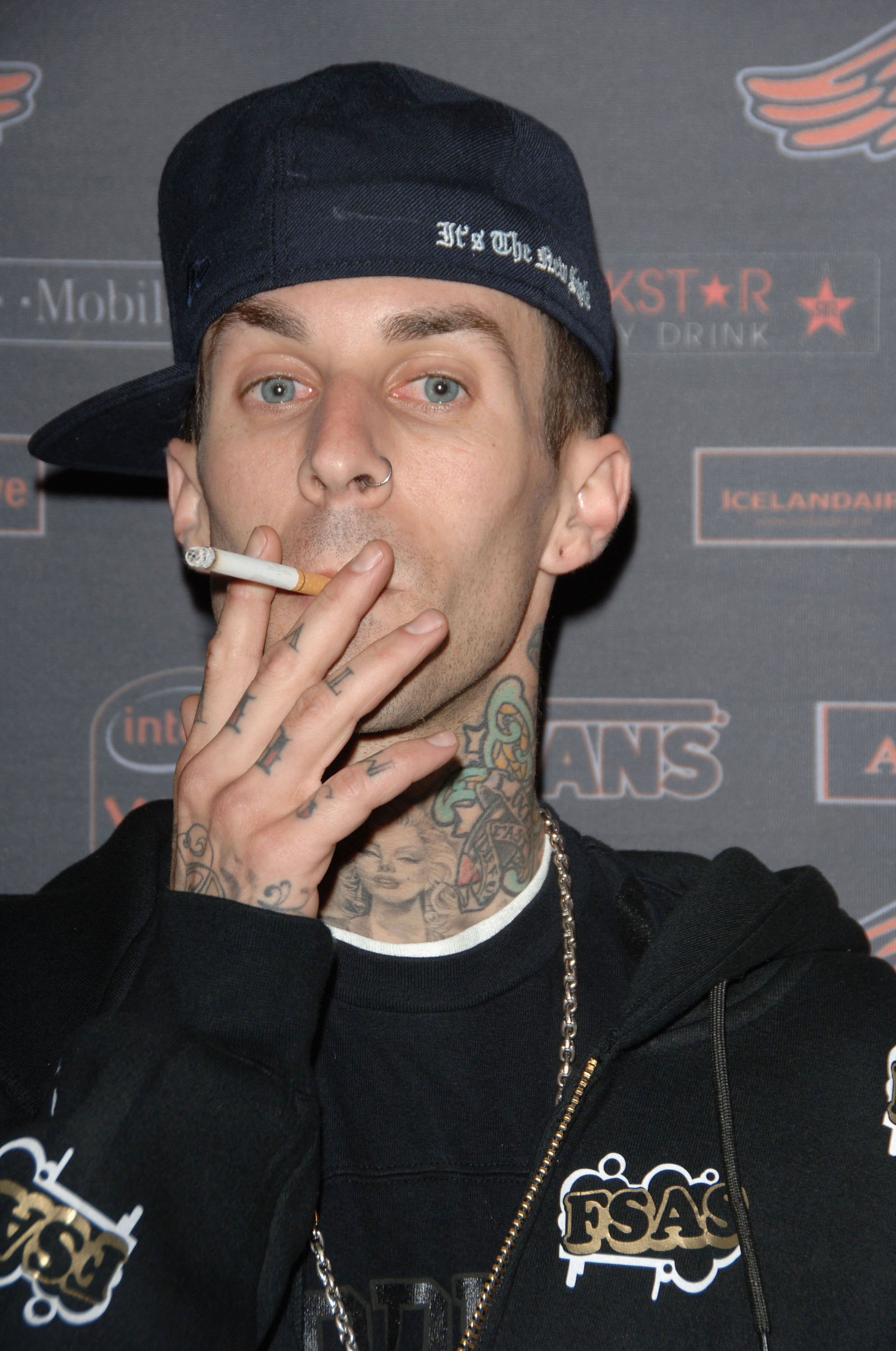Tattooed man in a backwards navy cap smoking a cigarette at a branded event backdrop, wearing a black FSAS hoodie and gold chain.