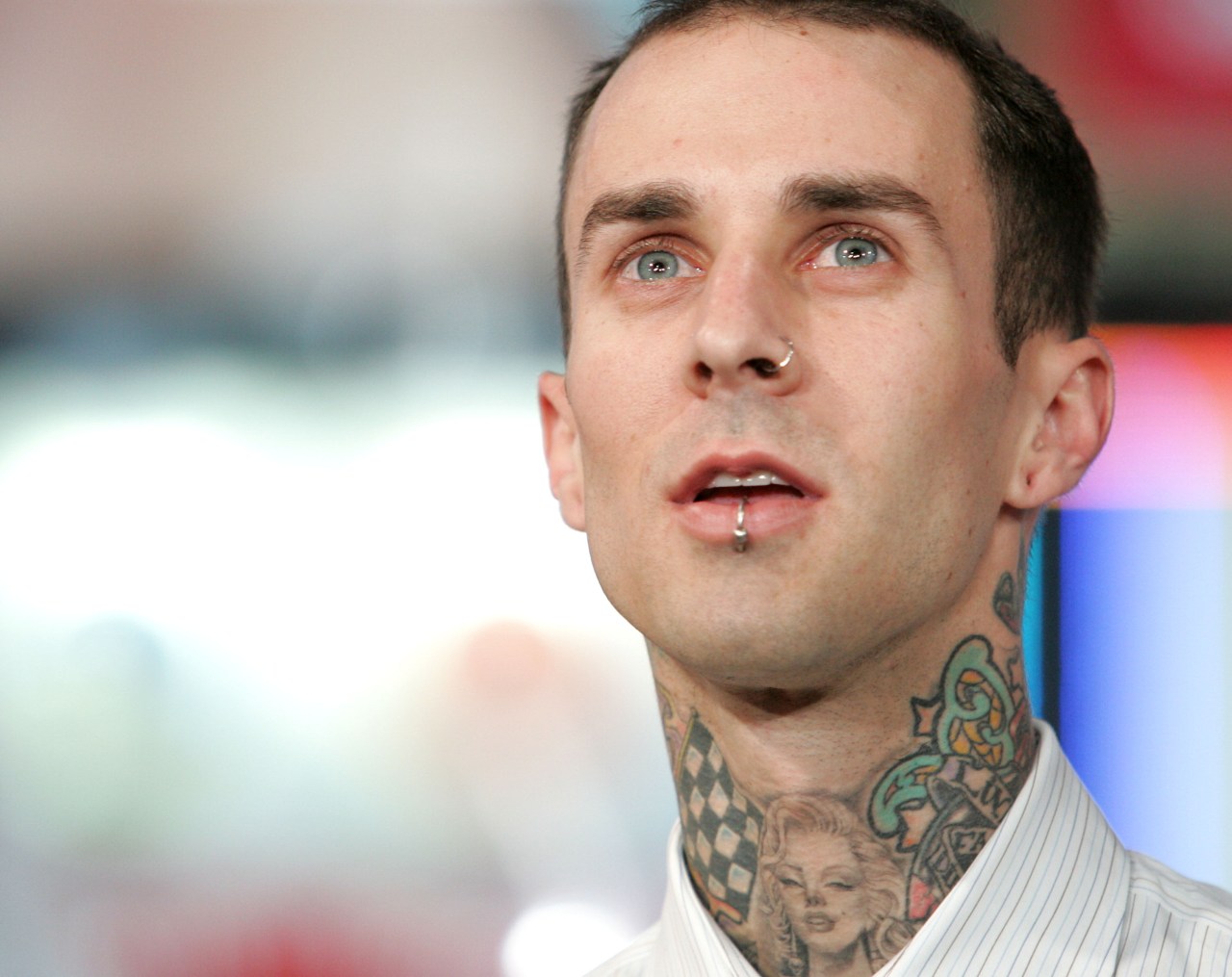 Close-up of a young man with blue eyes, nose and lip piercings, and extensive neck tattoos, looking upward against a blurred colorful background.