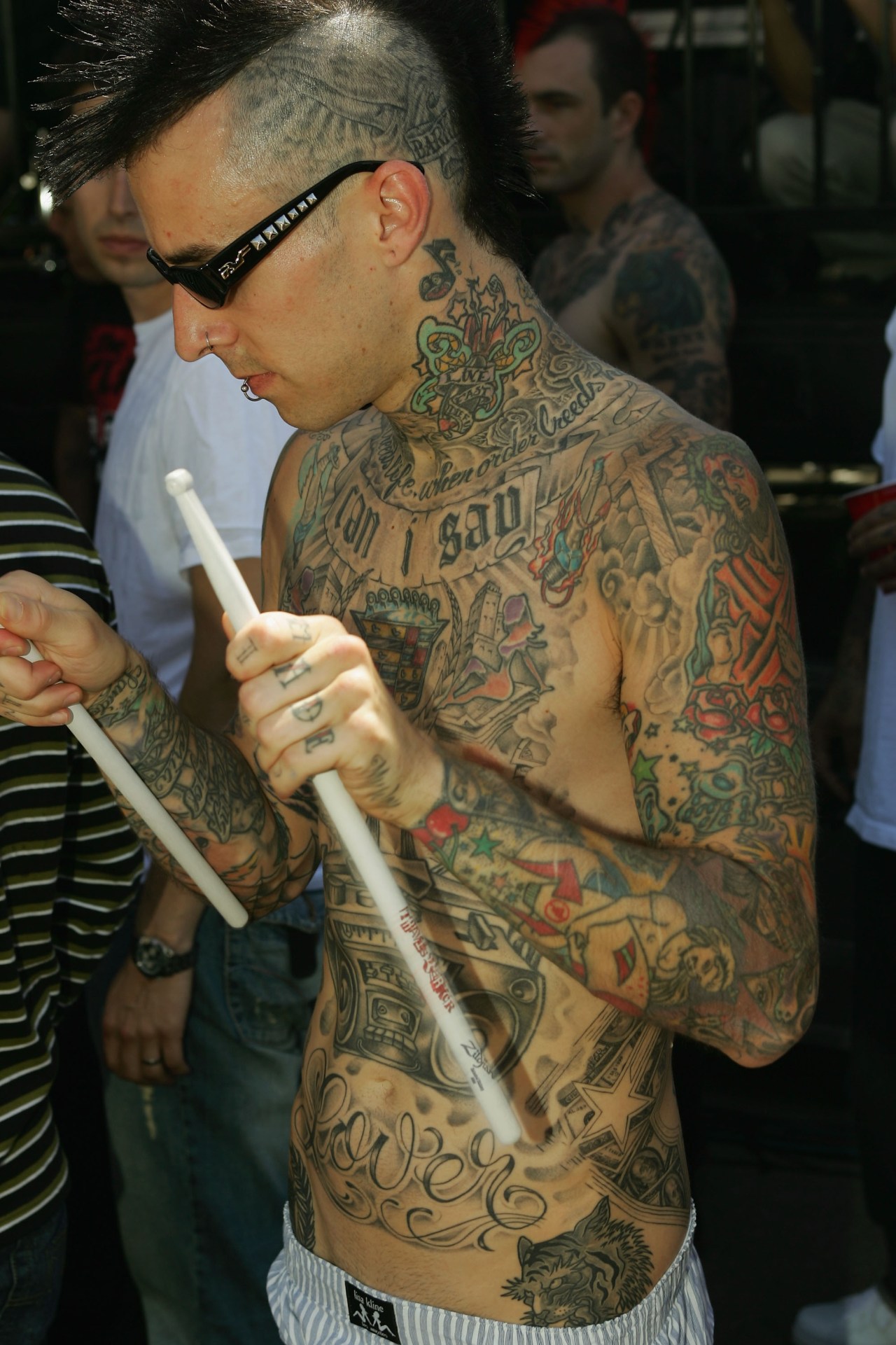 Shirtless tattooed man with sunglasses and a mohawk holding drumsticks backstage at an outdoor concert, full-body tattoos visible.
