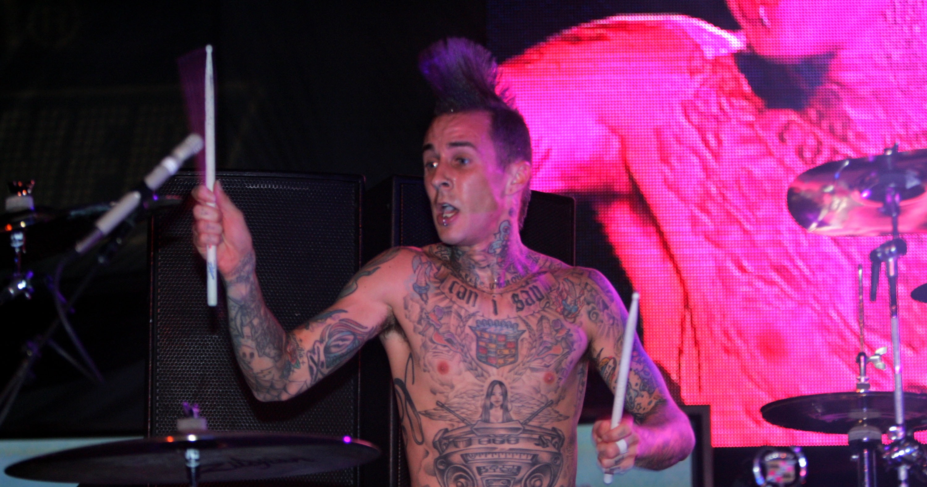 Shirtless, heavily tattooed drummer playing drums with drumsticks raised, performing on a concert stage with pink lighting.