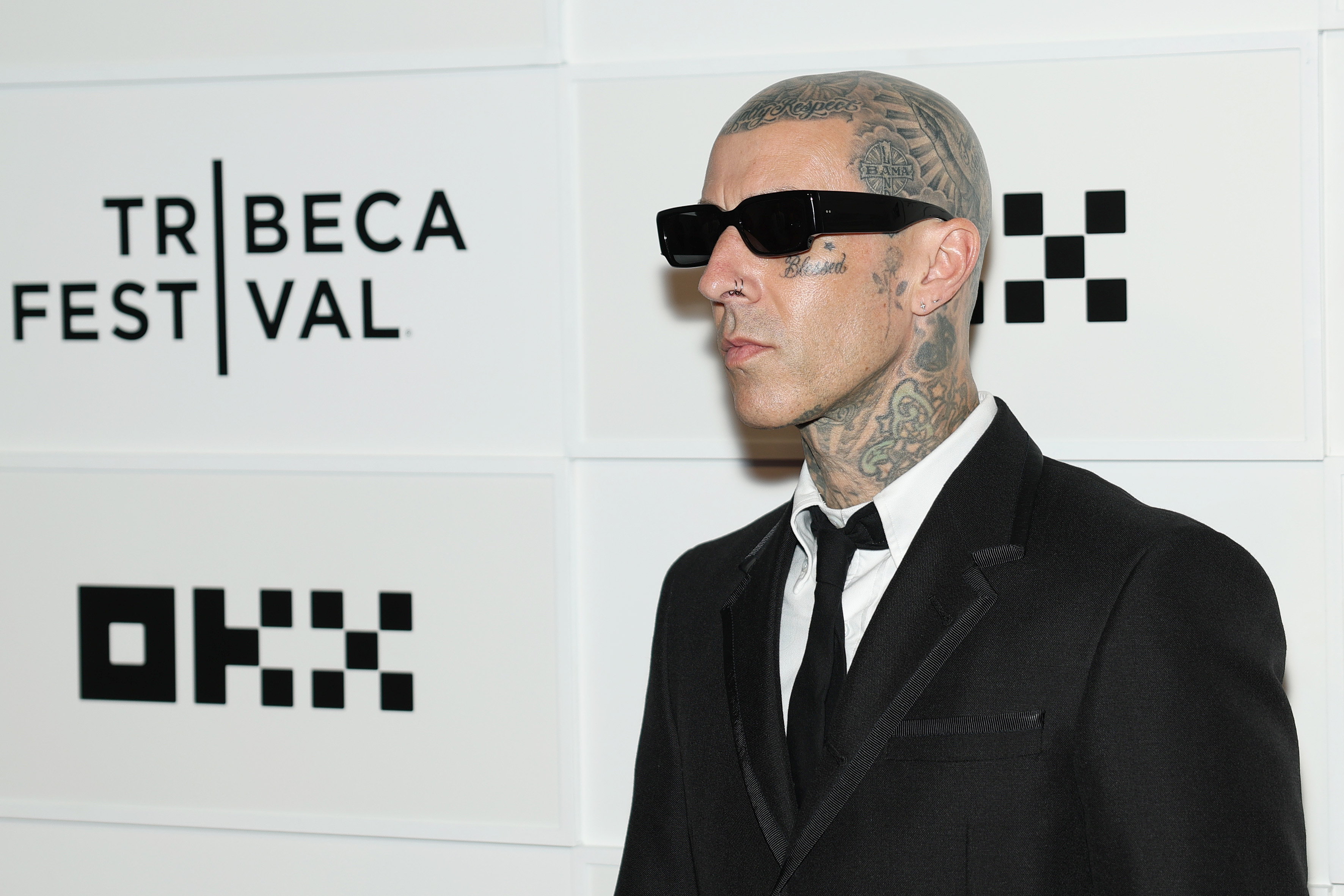 Side profile of a tattooed man in a black suit and dark sunglasses standing before a Tribeca Festival and OKX step-and-repeat wall.
