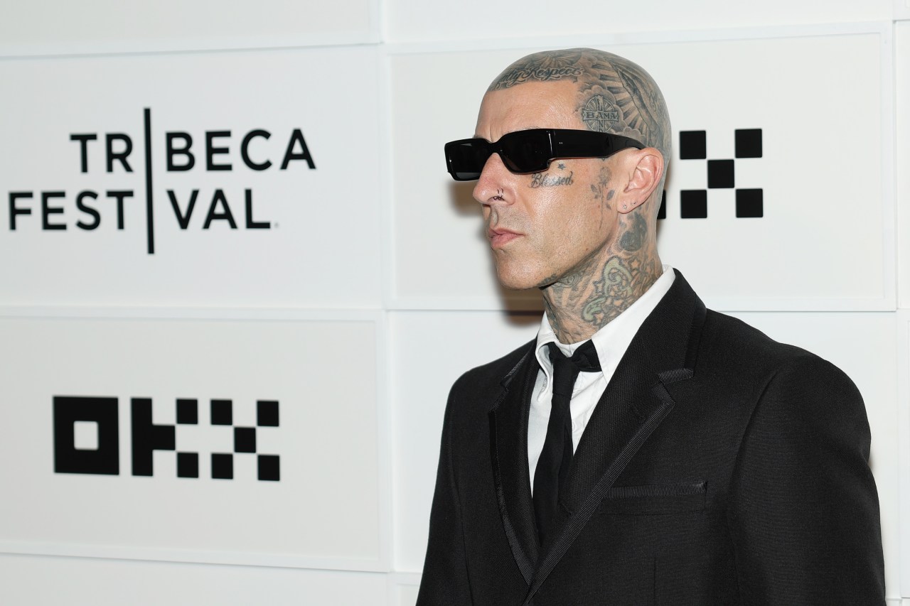 Side profile of a tattooed man in a black suit and dark sunglasses standing before a Tribeca Festival and OKX step-and-repeat wall.