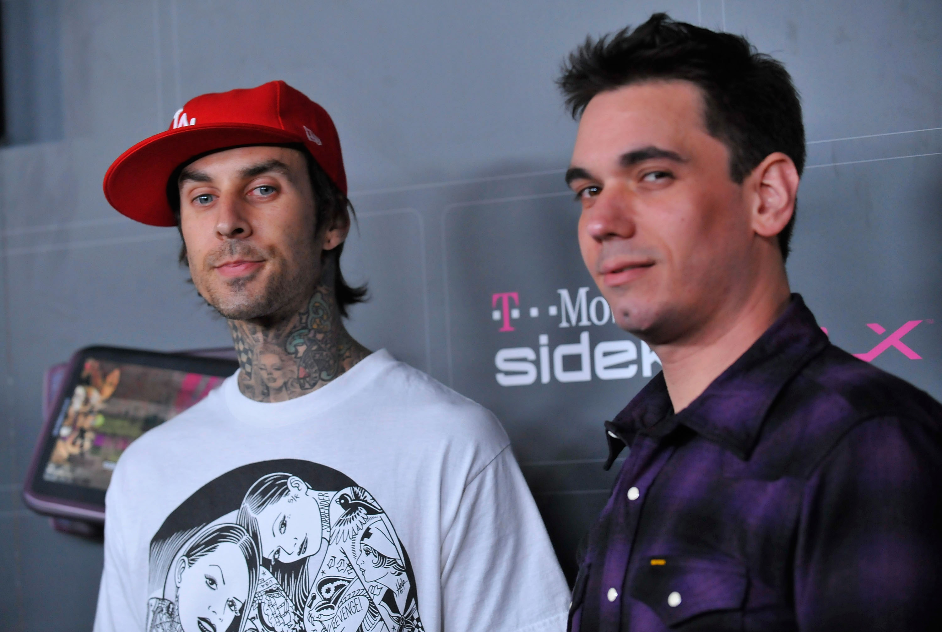 Two men stand in front of a T-Mobile Sidekick event backdrop; the man on the left wears a red LA cap and a graphic tee with visible neck tattoos.