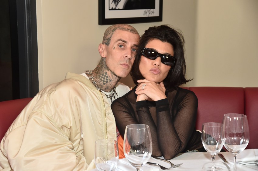 Heavily tattooed man in a cream bomber jacket sits beside a dark-haired woman in a black sheer top and dark sunglasses at a restaurant table.