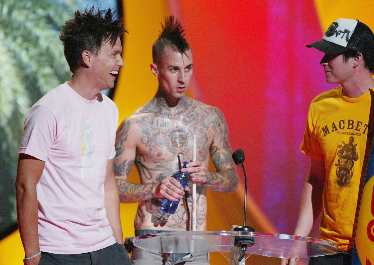 Three men on stage at an awards show — one in a pink tee, one shirtless with a red mohawk and full-body tattoos, one in a yellow tee and trucker hat — standing at a podium.