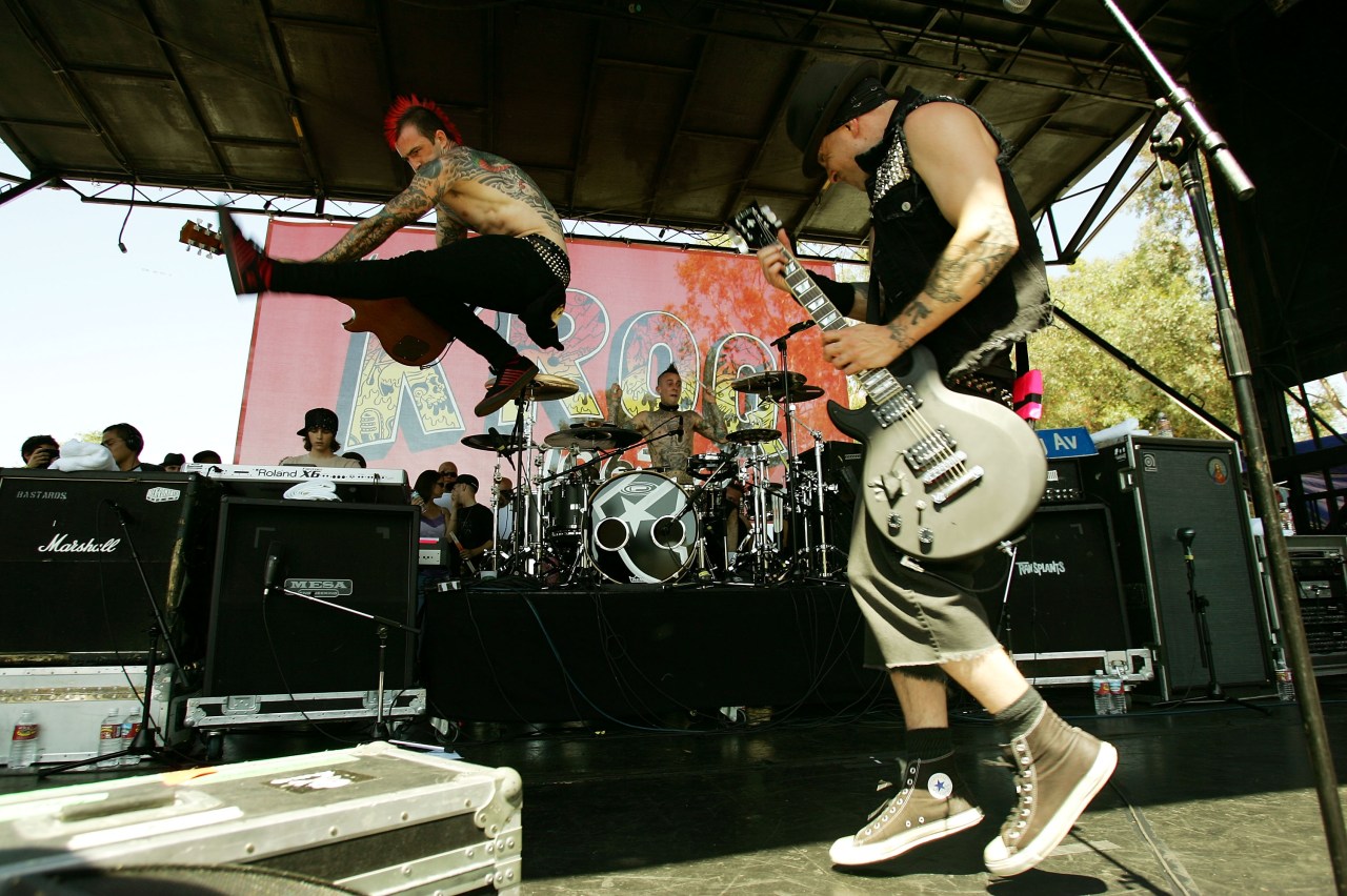 Shirtless tattooed man jumping in the air with a guitar while a drummer plays in the background and a guitarist performs at an outdoor stage.