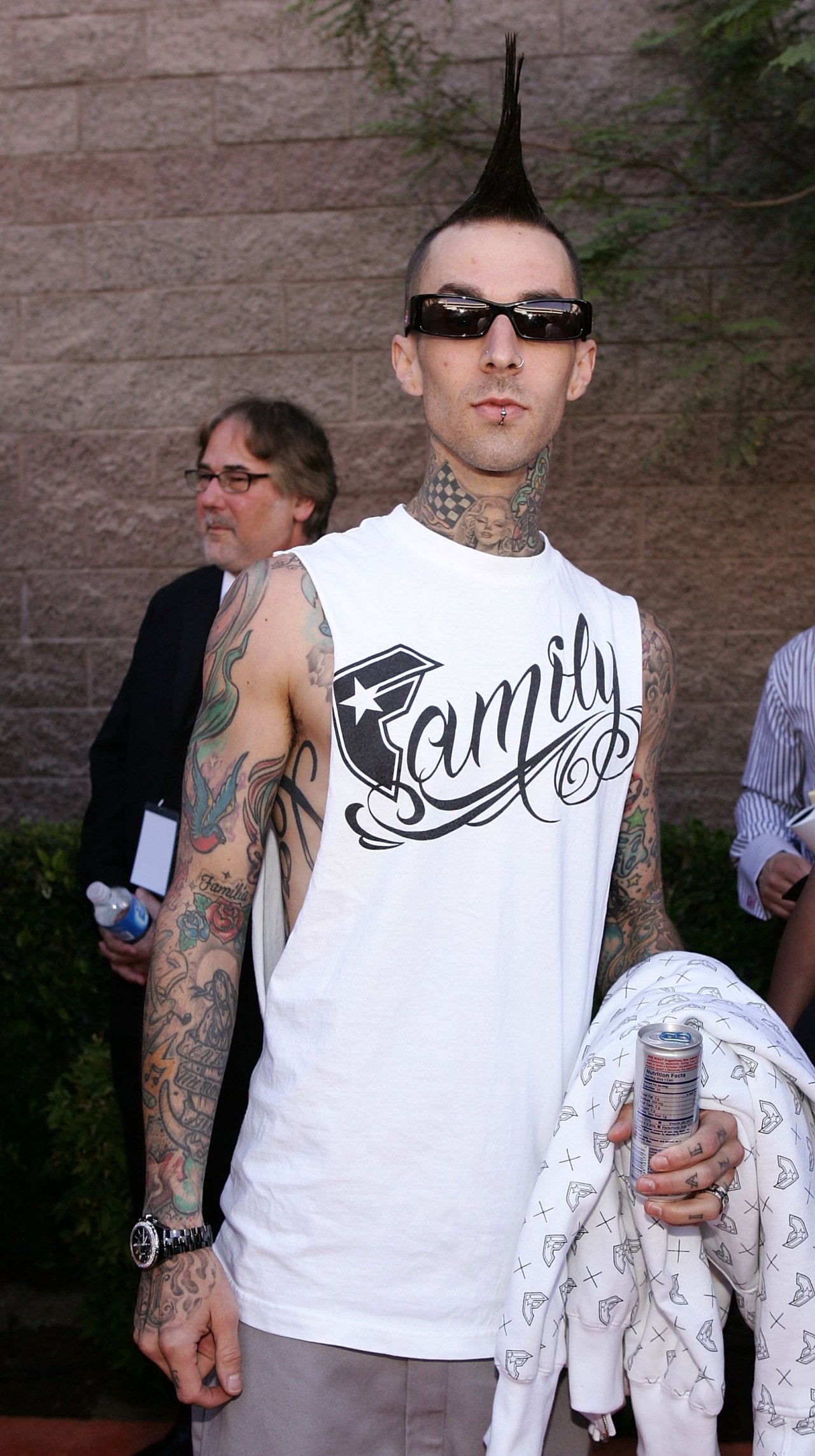 Tattooed man with a tall mohawk, dark sunglasses, and a white sleeveless shirt holding a beverage can outside an event venue.