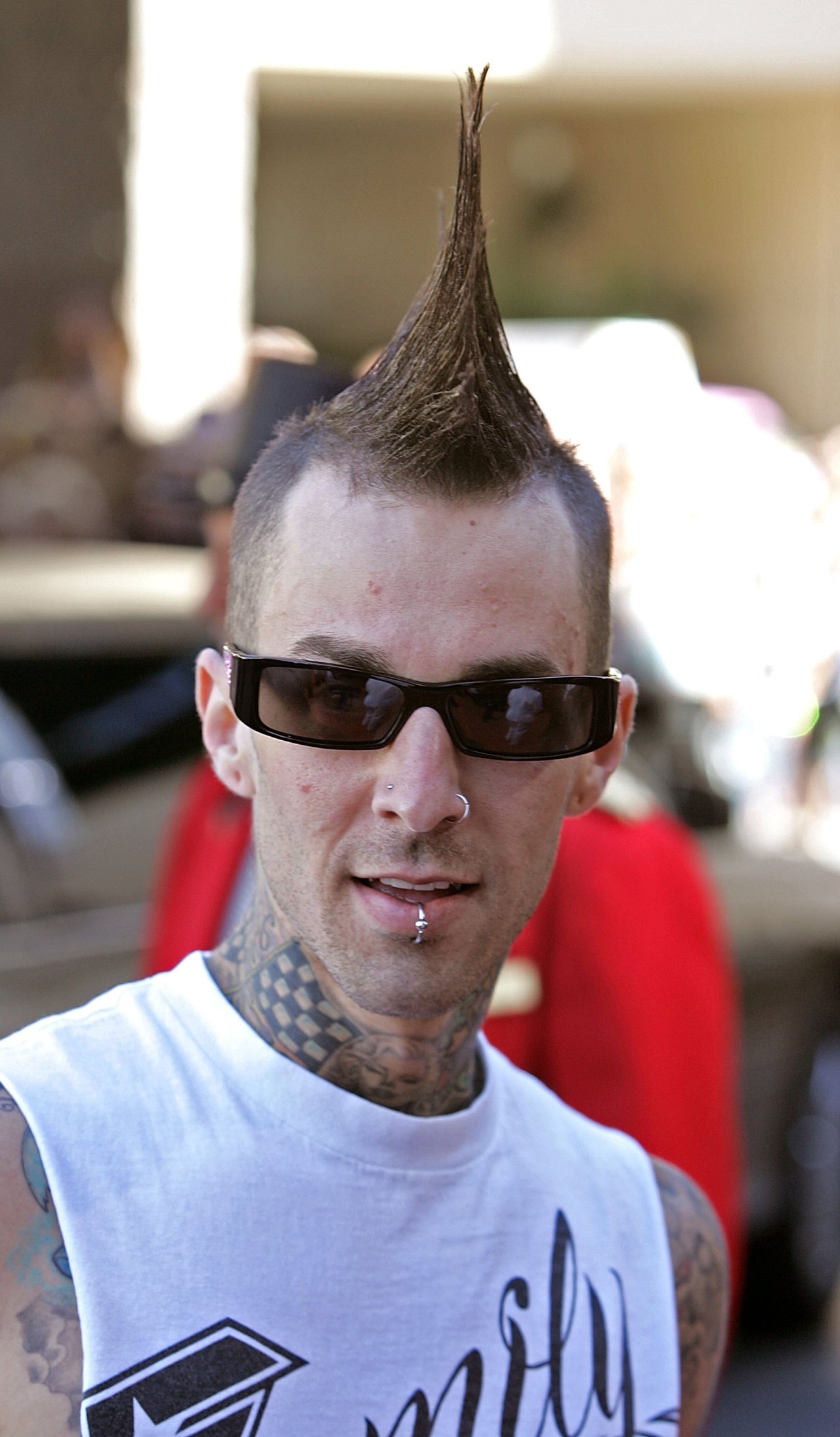 Close-up of a tattooed man with a tall mohawk, dark rectangular sunglasses, nose ring, and lip piercing wearing a white sleeveless shirt.