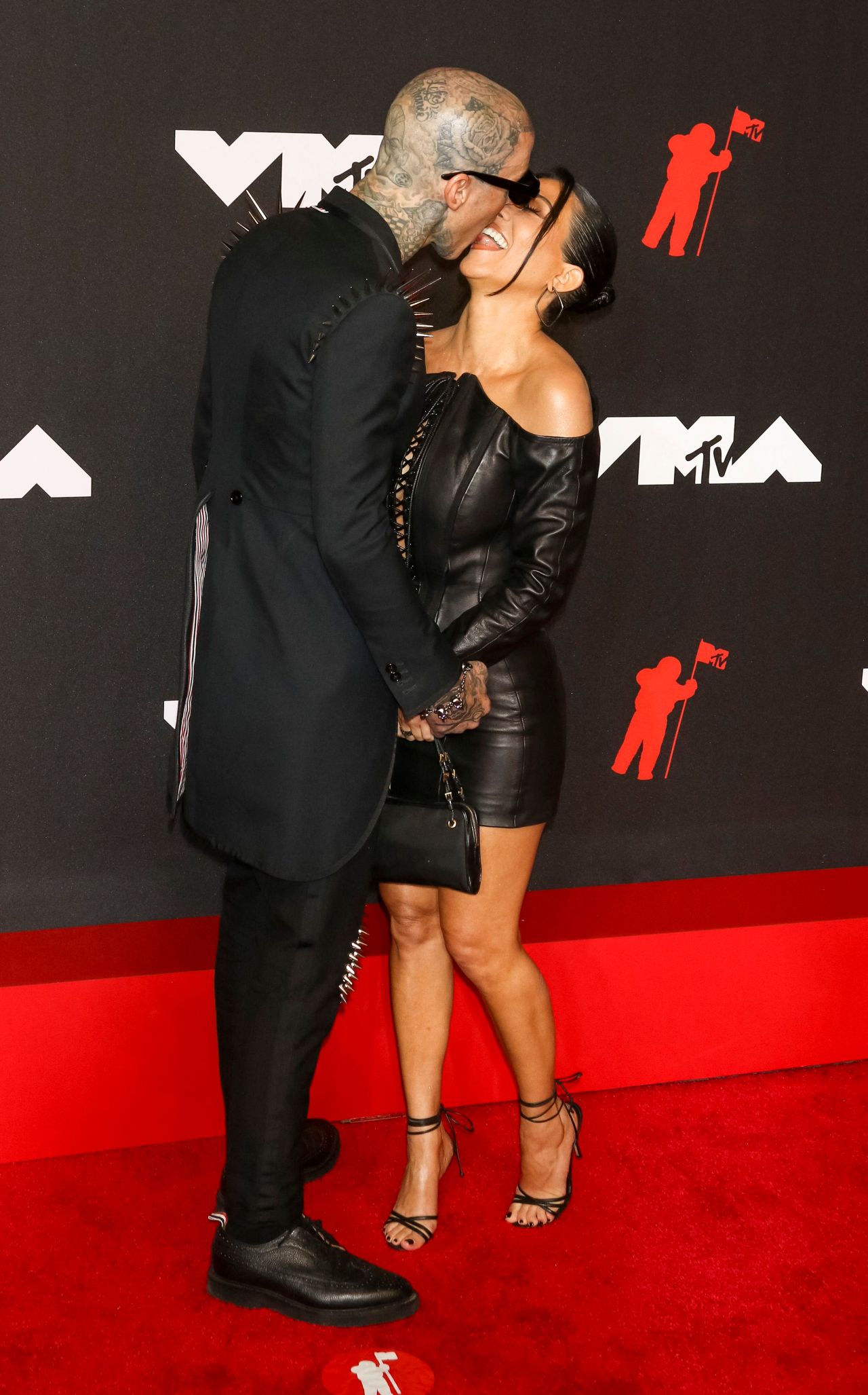 A heavily tattooed man in a black spiked blazer leans in to kiss a woman in a black leather mini dress on the MTV VMAs red carpet.