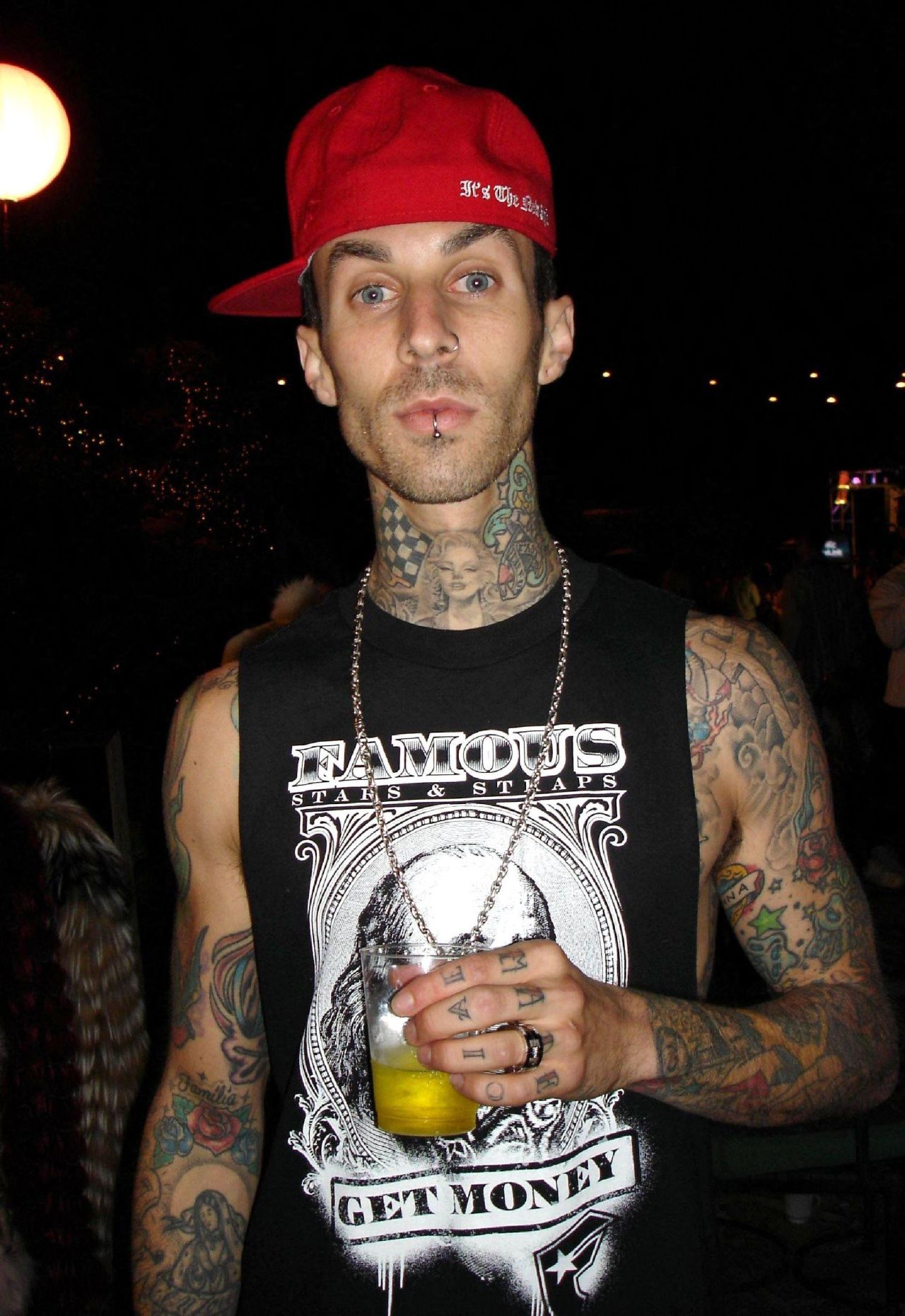 Man with extensive tattoos on arms and neck wearing a red snapback cap and black sleeveless Famous Stars & Straps shirt, holding a drink.