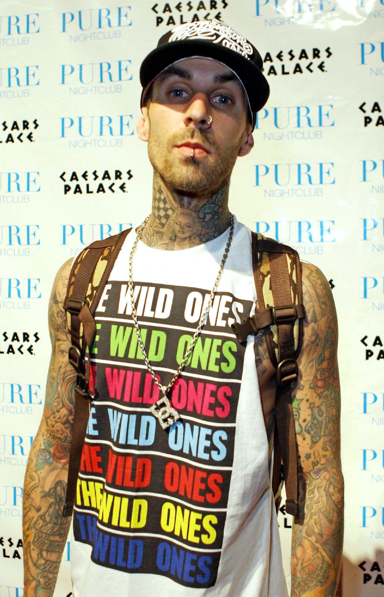 Heavily tattooed man in a black cap, 'The Wild Ones' tank top, and gold chain poses in front of a Pure Nightclub at Caesars Palace step-and-repeat backdrop.