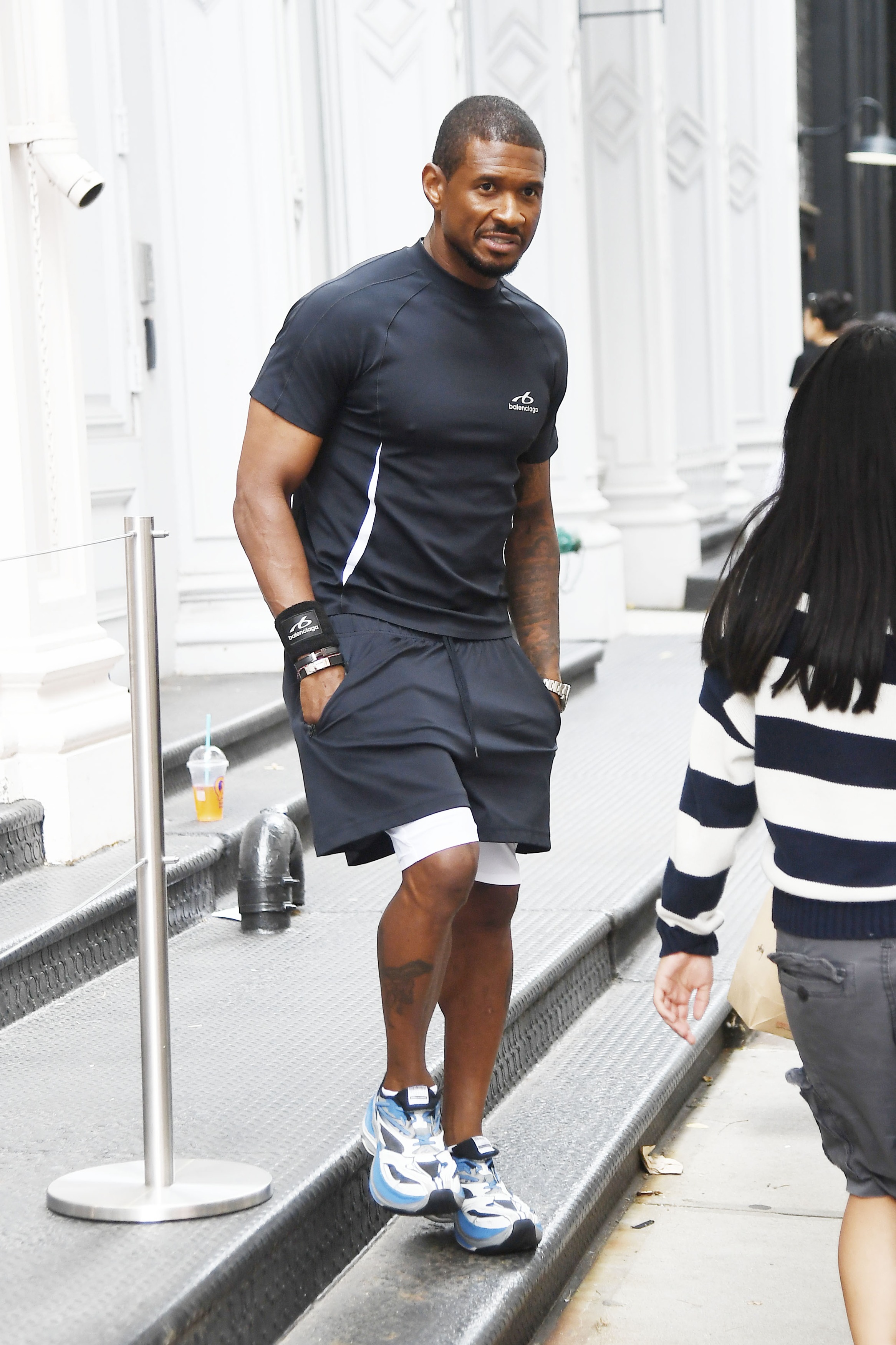 Usher walking through SoHo in a black Balenciaga workout outfit with layered shorts, wristbands, and blue-and-white sneakers