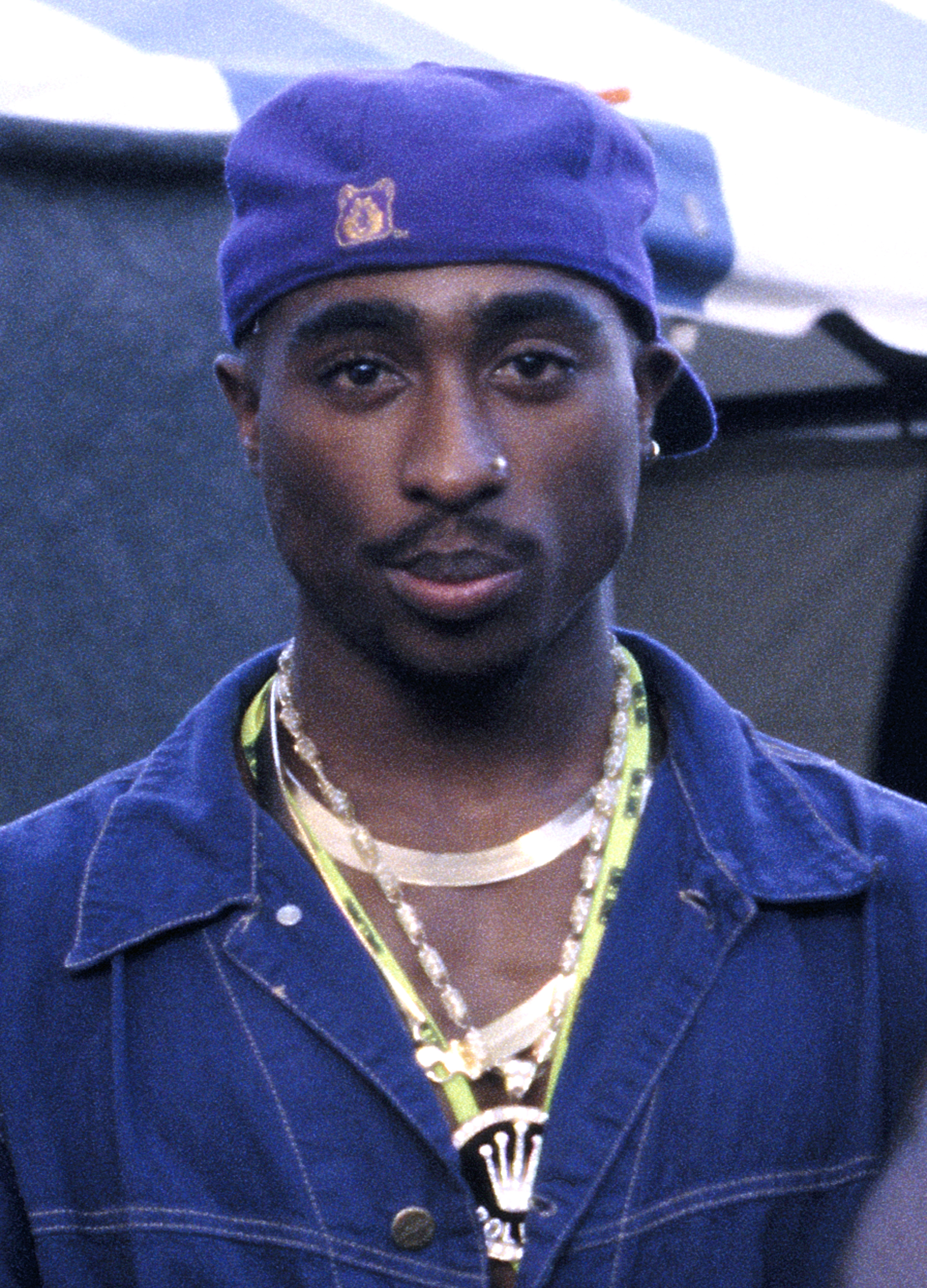 Tupac Shakur wearing a purple cap and blue denim jacket with gold chains backstage at KMEL Summer Jam 1992.