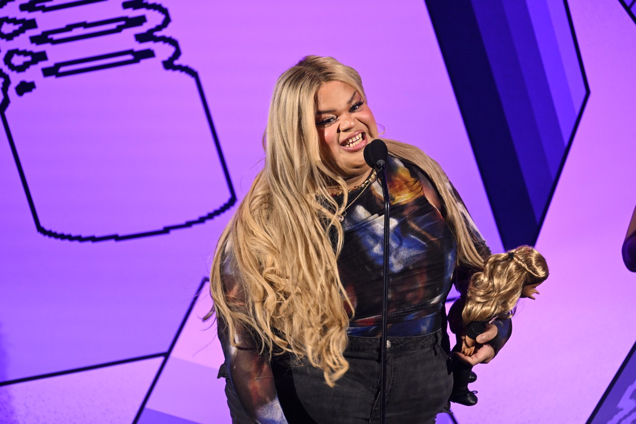 Jools Lebron with long blonde hair speaks at a microphone on stage at the Webby Awards, holding a small blonde doll, against a purple pixelated backdrop.