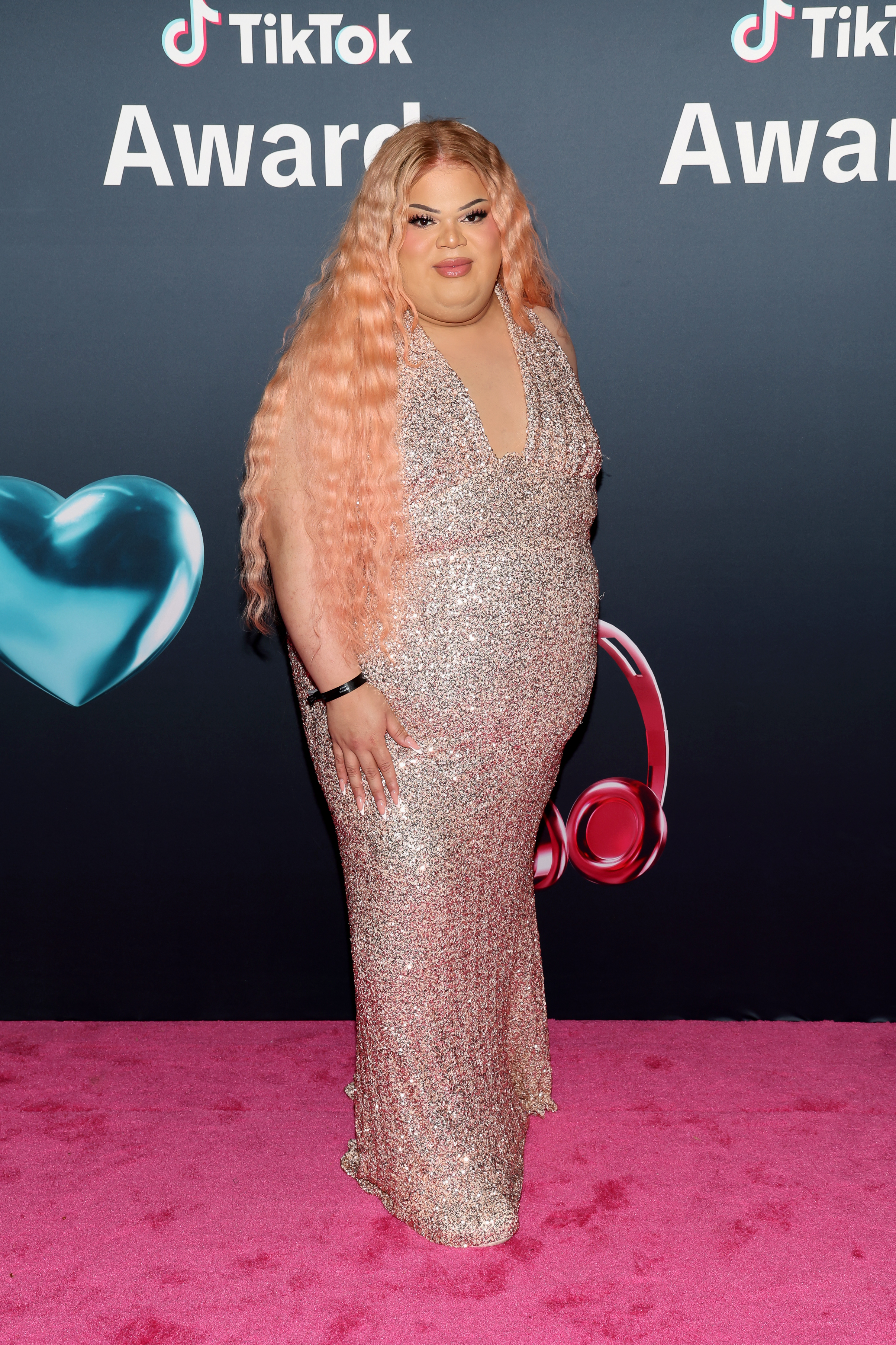 Jools Lebron in a floor-length sequined gown with long wavy pink hair, posing on a pink carpet in front of a TikTok Awards backdrop.