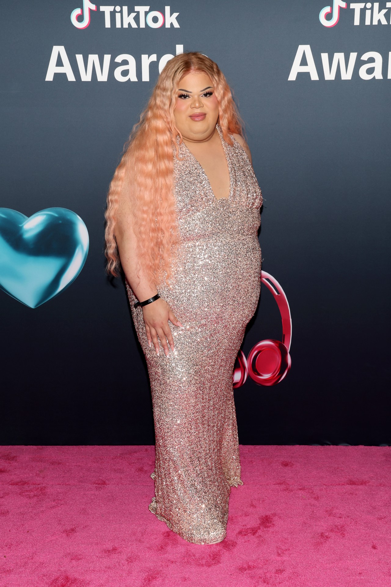 Jools Lebron in a floor-length sequined gown with long wavy pink hair, posing on a pink carpet in front of a TikTok Awards backdrop.