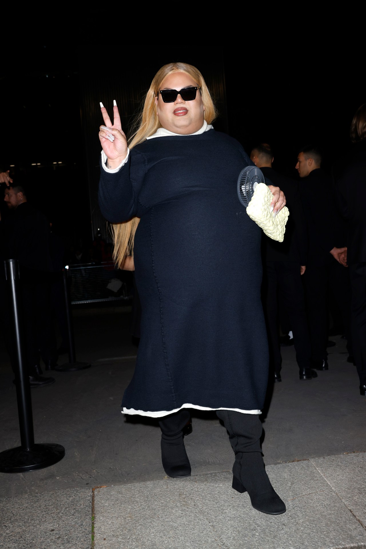 Person with long blonde hair in a navy midi dress and black sunglasses making a peace sign outside a fashion show venue.