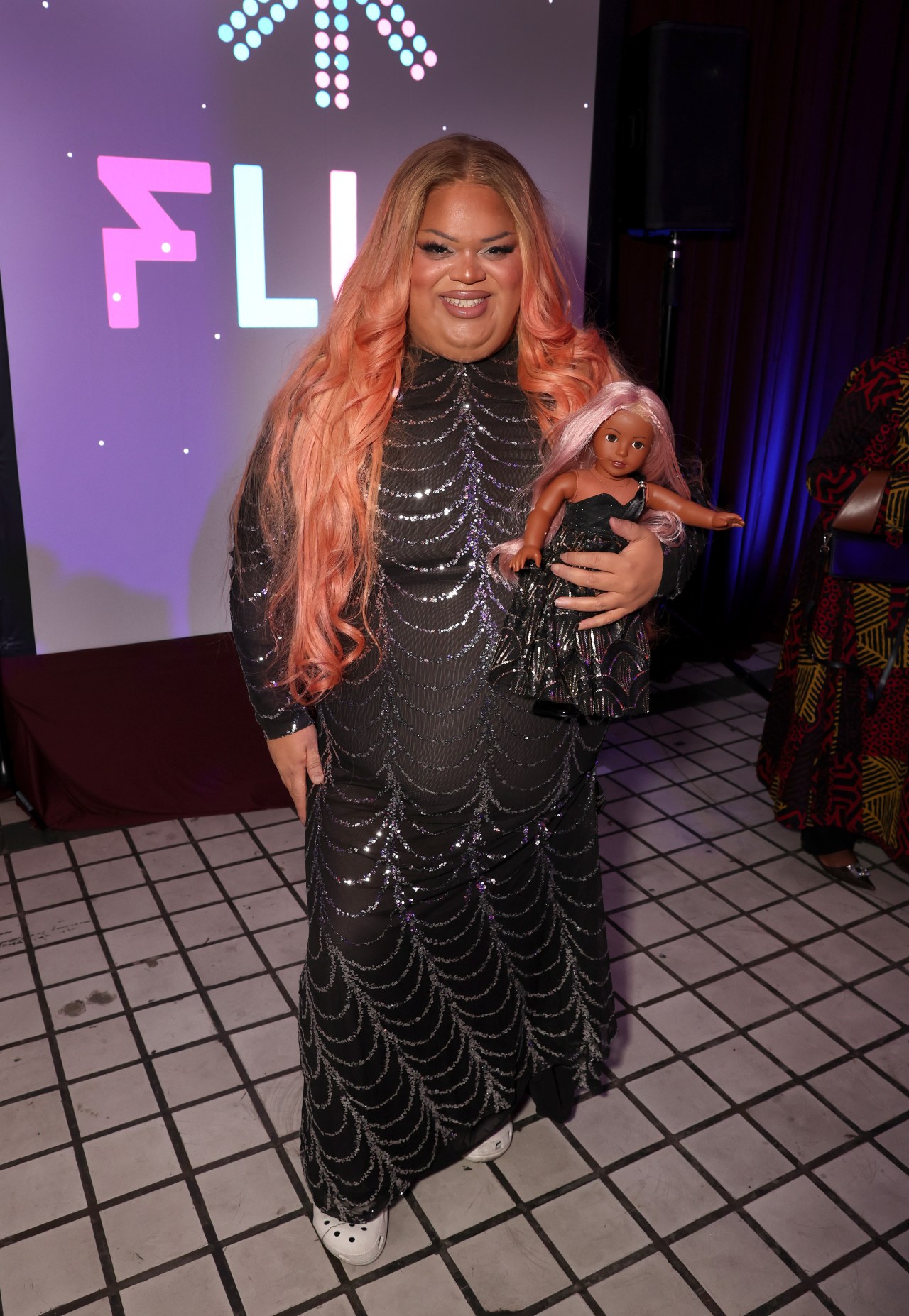 Person with long pink-orange hair in a black sequined gown smiling and holding a doll with matching pink hair and dress at an event.