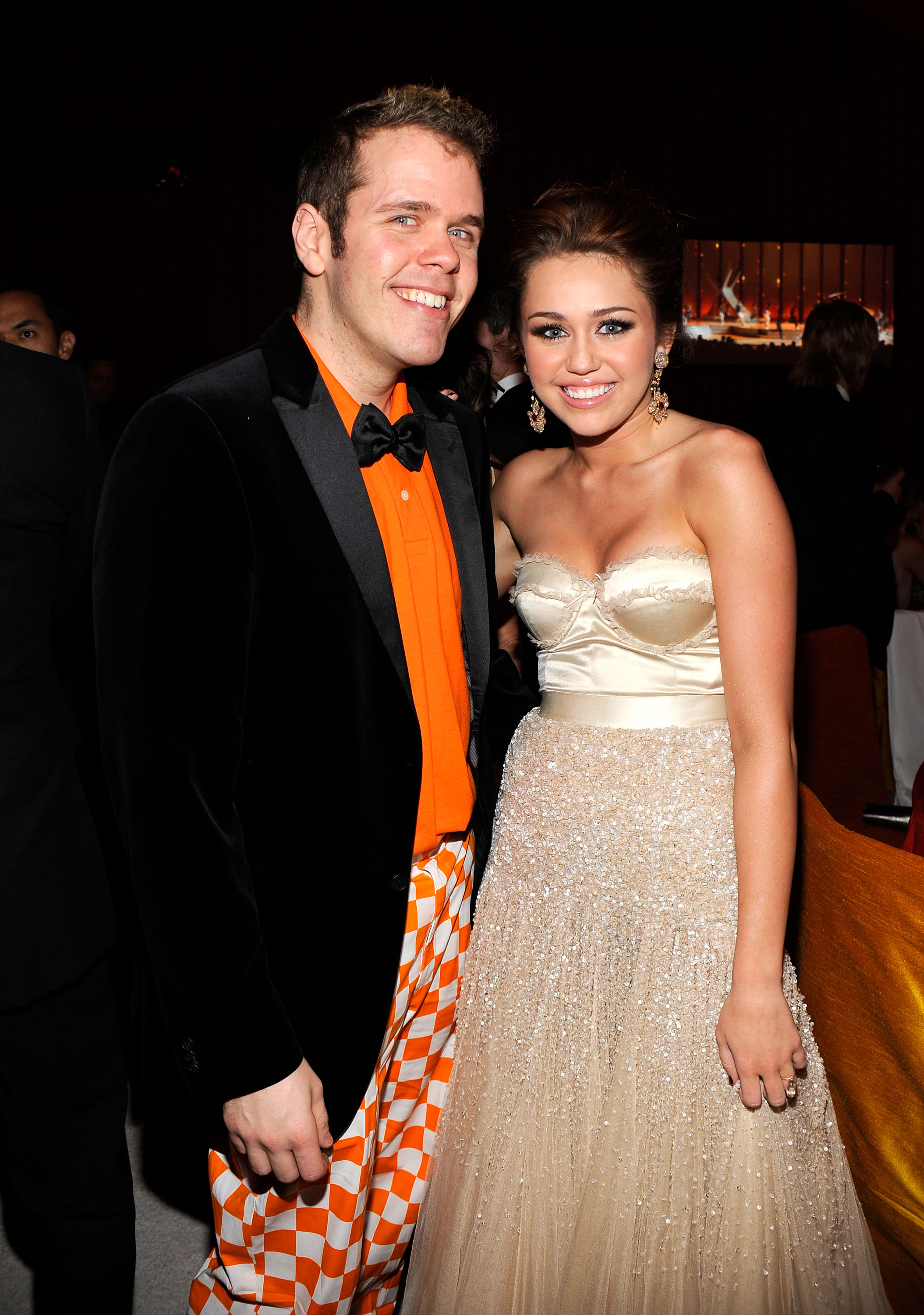 Perez Hilton in an orange shirt and checkered pants stands beside a young woman in a strapless beaded gown at a formal event.