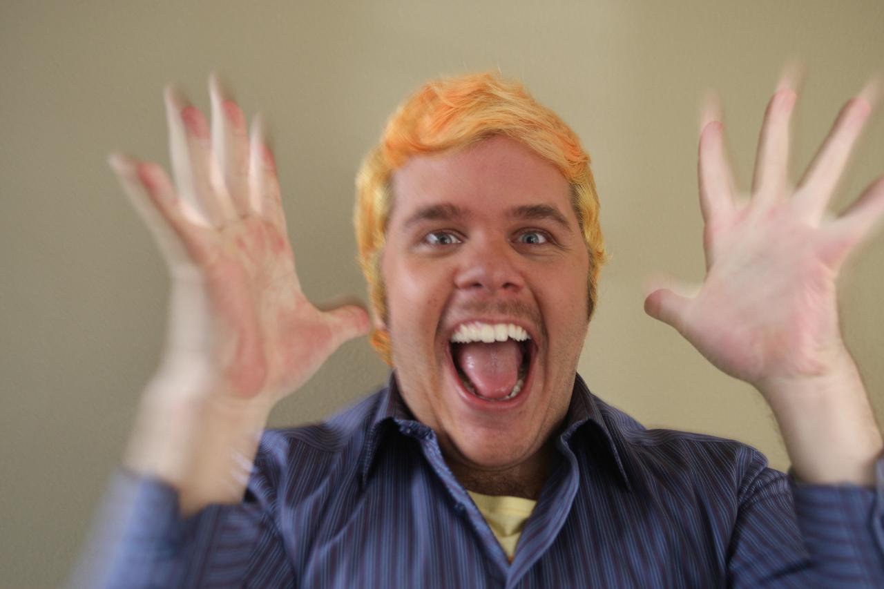 Man with bright orange hair wearing a blue striped shirt, mouth wide open and both hands raised toward the camera.