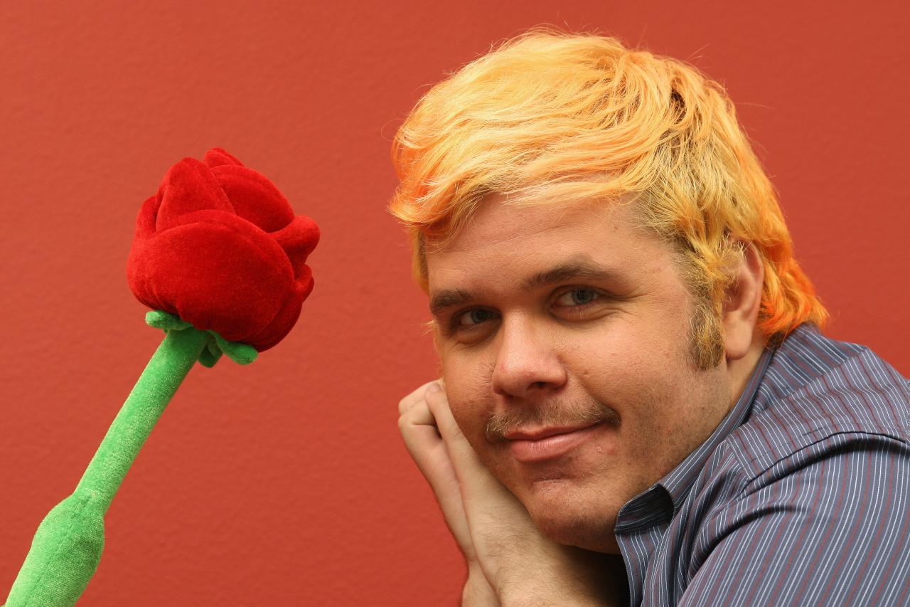 Man with orange dyed hair resting chin on hand, smiling beside a large stuffed red rose against a red wall.