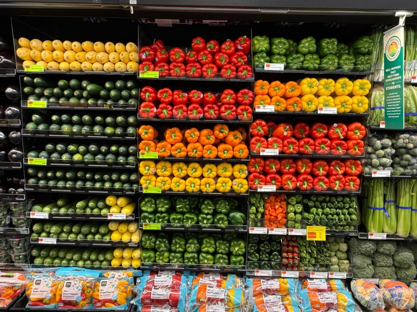 Grocery store produce aisle with shelves of red, orange, yellow, and green bell peppers, squash, and other vegetables at a Whole Foods Market.