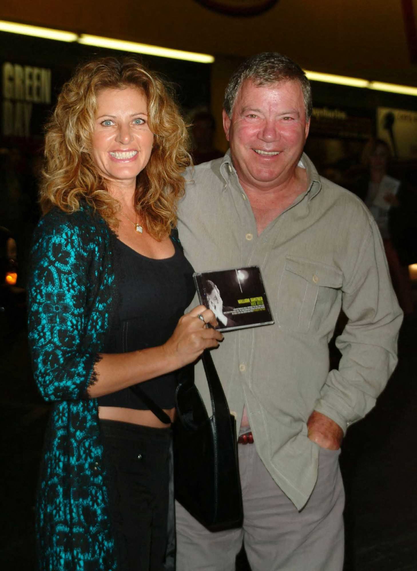 William Shatner and a woman with curly blonde hair smile together, holding a CD, inside a record store.