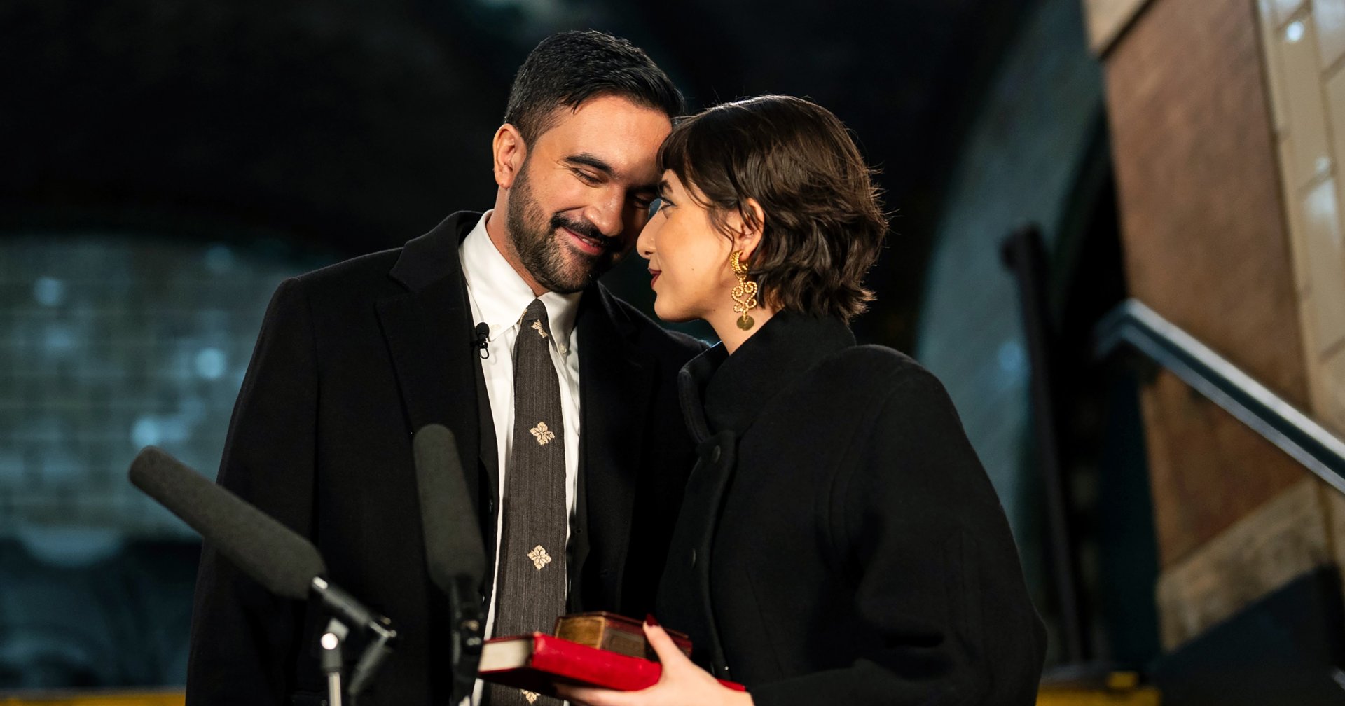 Zohran Mamdani and Rama Duwaji lean their heads together and smile at each other in a subway station, she holding a small red book.